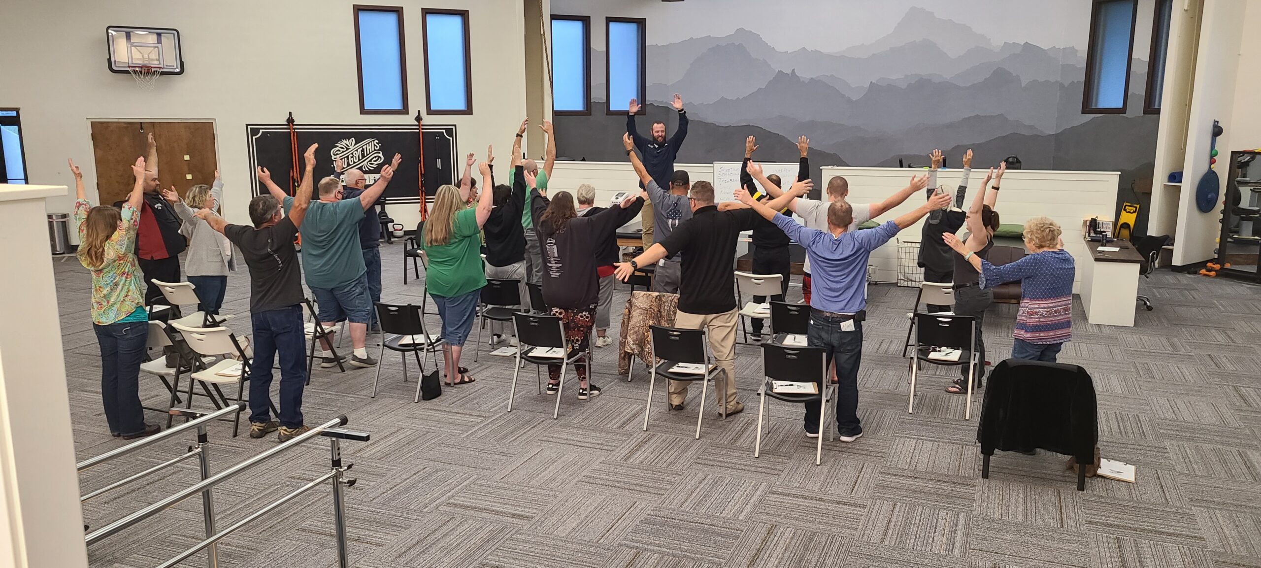 A community workshop in the clinic: attendees stand with their arms raised while the therapist leads