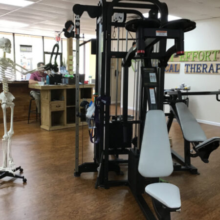A strength-training machine in the clinic exercise area, with a model skeleton and the front desk behind it