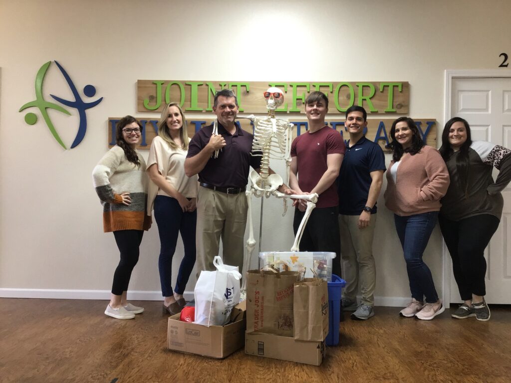 Seven team members standing beside an anatomical skeleton under the Joint Effort Physical Therapy sign
