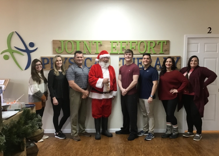 Team members and a person dressed as Santa Claus posing under the Joint Effort Physical Therapy sign