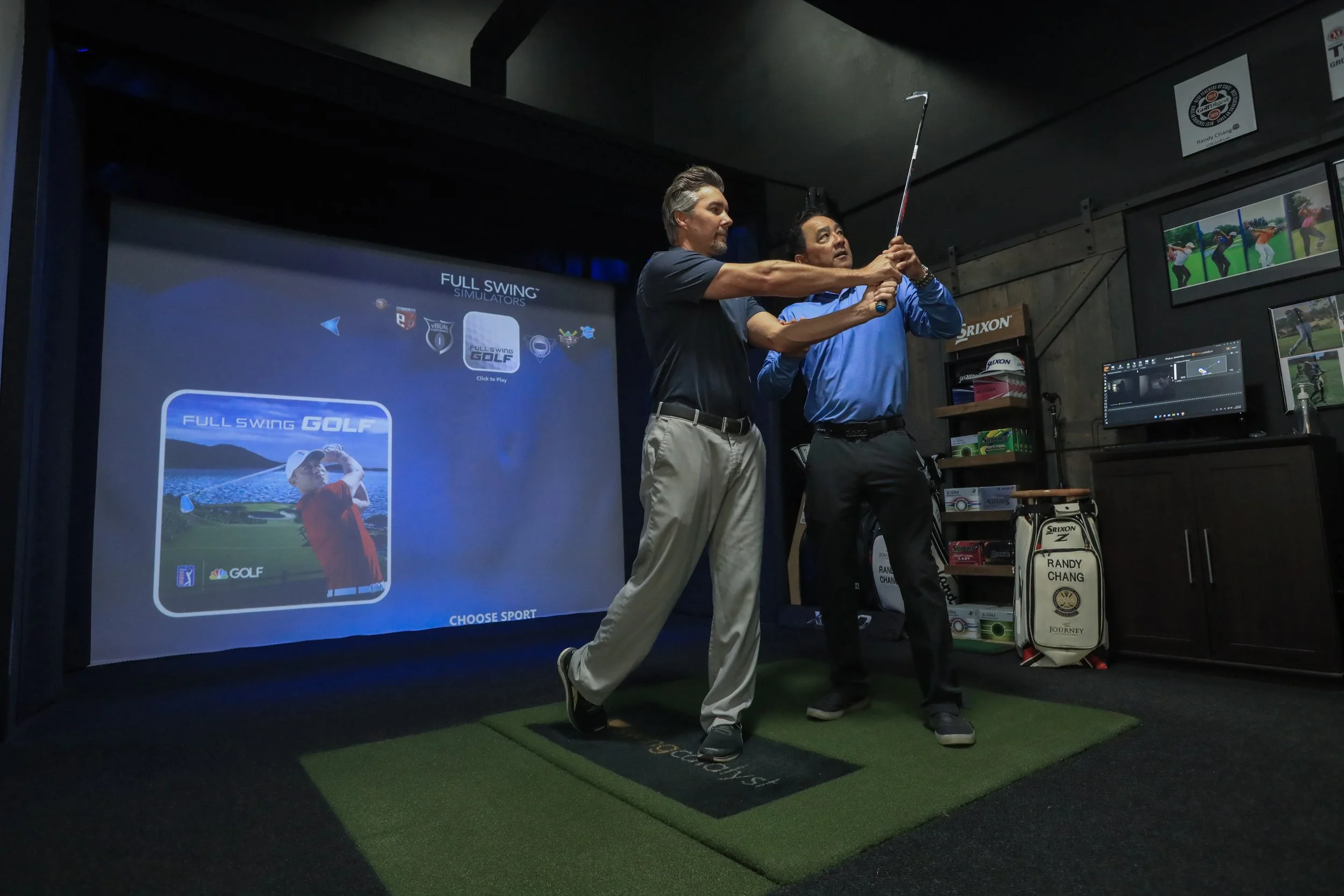 Two men at a golf simulator screen, one showing the other how to hold and swing a club