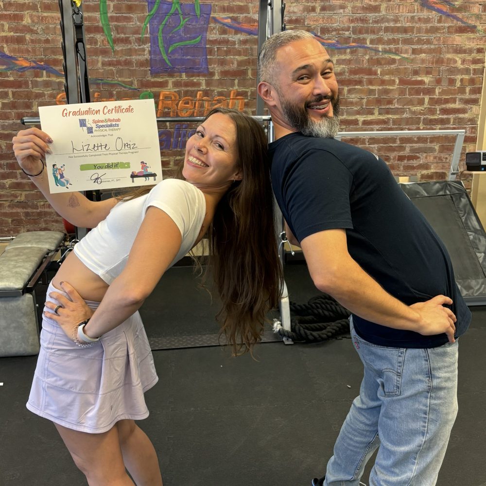 Two people laughing and posing with a certificate in front of a brick wall